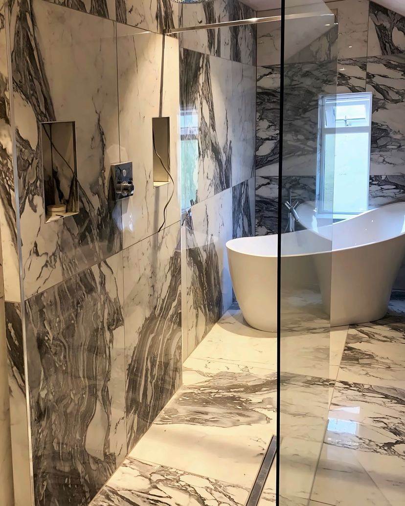 Walk-in shower with tiled niches in the same marble-effect bathroom, the bath beyond the glass